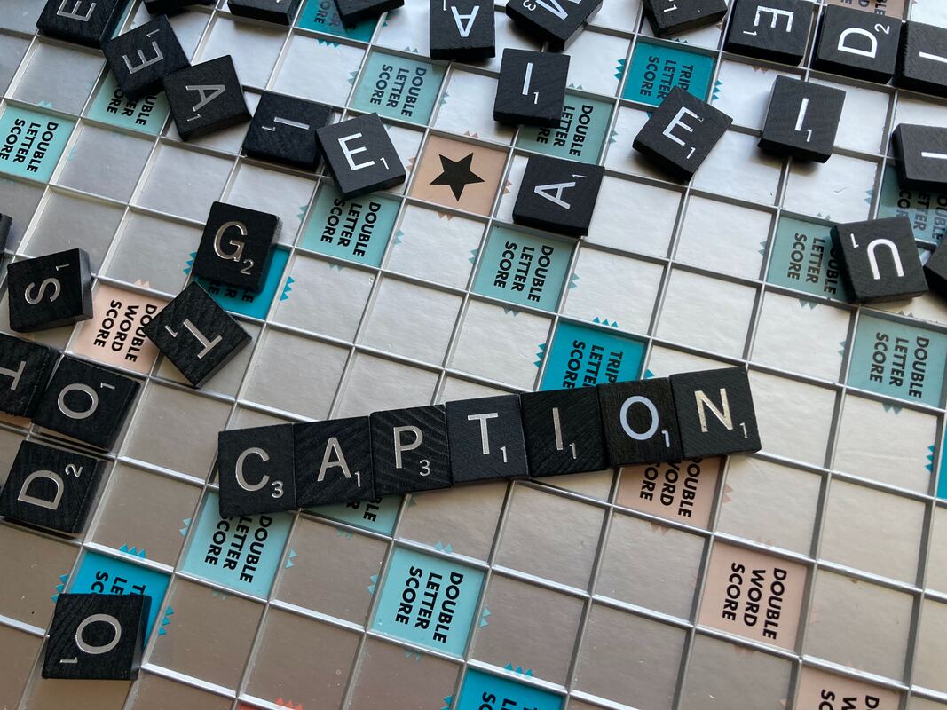 A Scrabble board with various black tiles with white letters scattered around. In the center, the word caption is spelled out with Scrabble tiles.