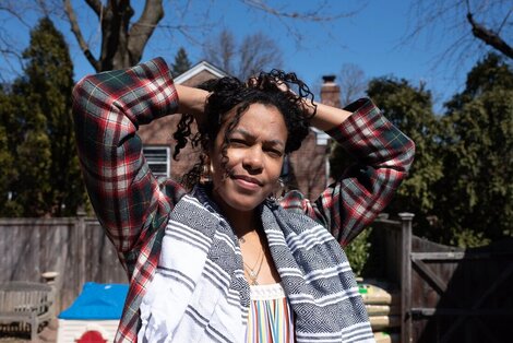 Dra. Raina Leon with her hands in her curly black hair, in the backyard of her home; in a flannel and scarf. 